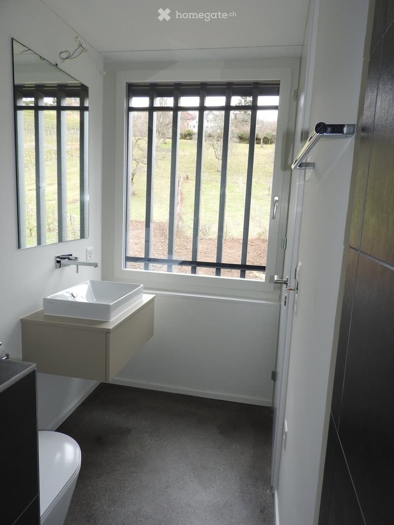 Modern bathroom with white walls, grey floor, large window with metal bars, sink with faucet, mirror, and toilet.