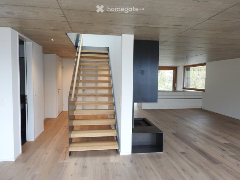 Modern hallway with wooden staircase, polished wooden floors, white walls, and recessed lighting. Features a black fireplace and a view into a kitchen area with white cabinets.