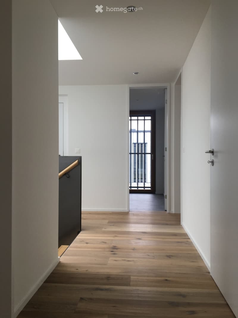 A modern hallway with light wooden flooring, white walls, and recessed ceiling lighting. A staircase with a dark railing is visible on the left, and a doorway leads to another room with a glass door and metal railing.