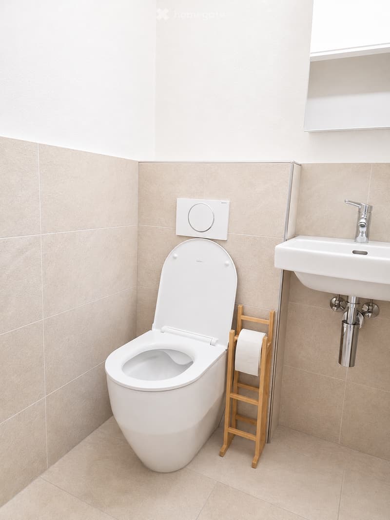 Modern bathroom with a white toilet, a sink with a chrome faucet, and a wooden toilet paper holder. The walls are tiled in a light beige color, and the floor is covered with matching tiles.