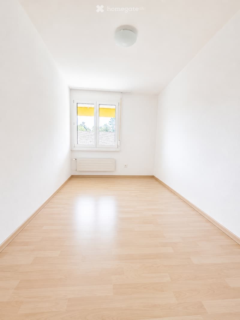 Empty room with light wooden flooring, white walls, a single window with blinds, and a radiator below the window.