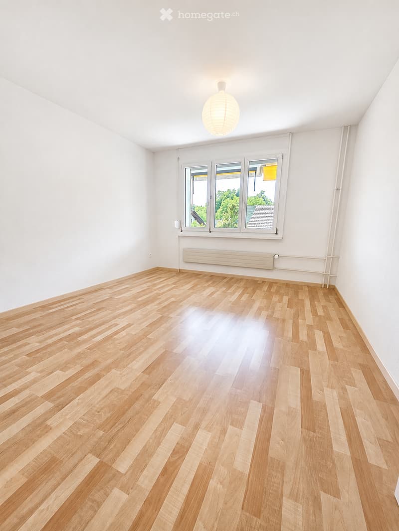 Empty room with light wooden flooring, white walls, large windows, and a single hanging light fixture.