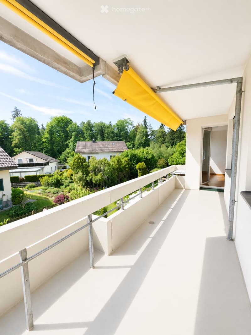 Balcony with white flooring, metal railing, and a yellow retractable awning, overlooking a green garden and neighboring houses.