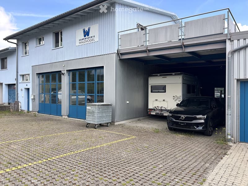 Two-story industrial-style building with a large garage entrance, featuring a camper van and a black car inside, along with a sign reading "CARROSSERIE WALDVOGEL".
