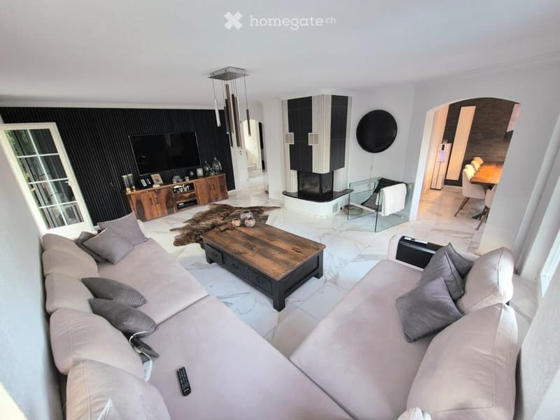 Modern living room with a white marble floor, a large sectional sofa, a wooden coffee table, a fireplace, a TV, and a dining area visible in the background.