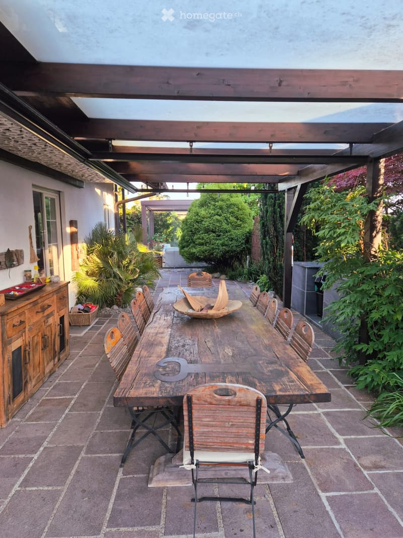 An outdoor patio with a wooden table and chairs, surrounded by lush greenery and a covered wooden structure.