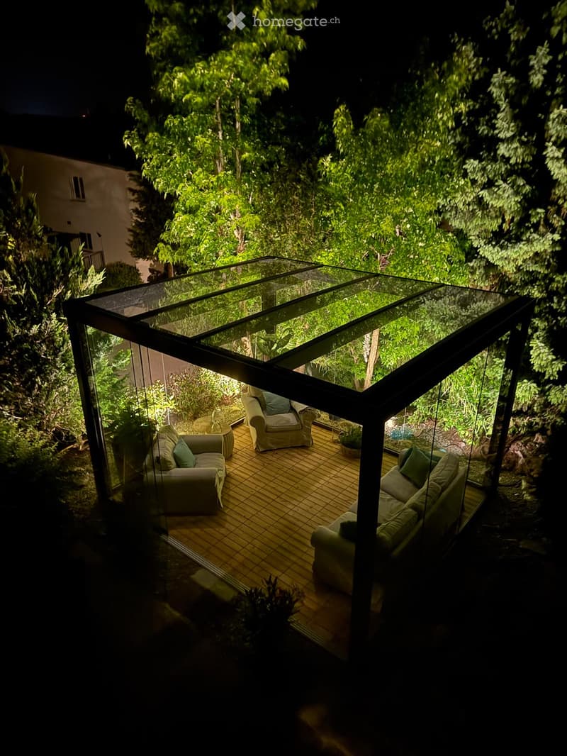A modern glass-enclosed outdoor seating area with wooden flooring, featuring a sofa, chairs, and a small table surrounded by lush greenery.