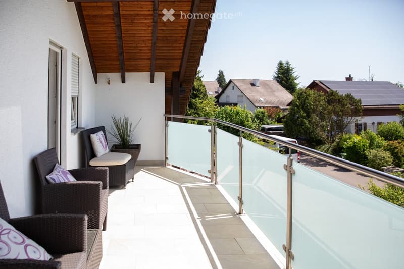 Balcony with modern glass railing, wicker chairs, and a small sofa, overlooking a residential area with trees and houses.