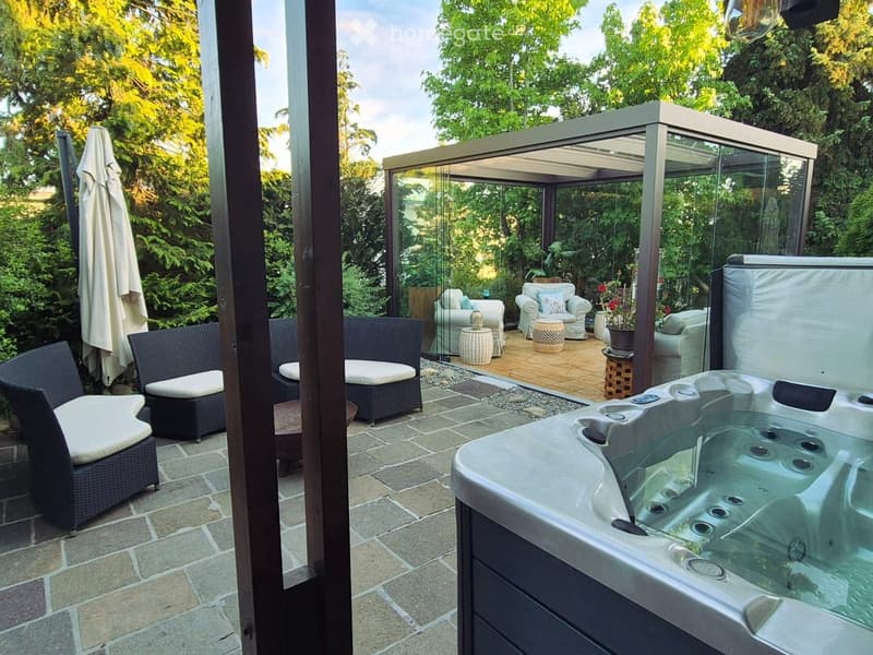 A well-maintained outdoor patio area with a hot tub, several seating options including wicker chairs and sofas, and a covered pergola with glass walls. The space is surrounded by lush greenery and features a stone-tiled floor.