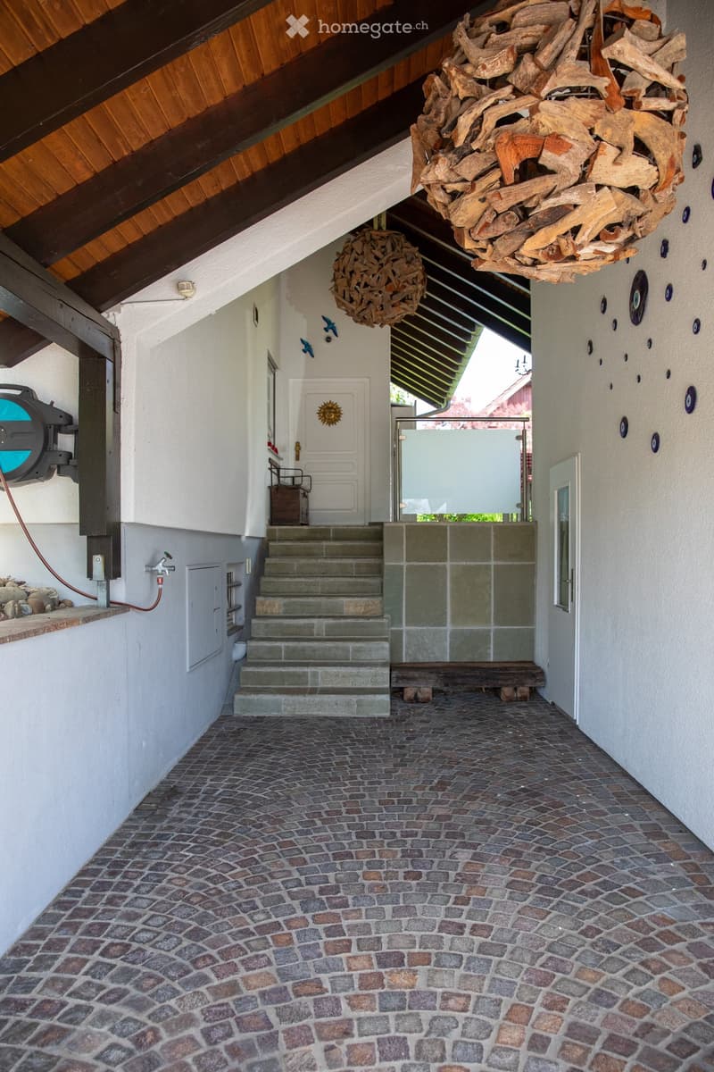 A spacious hallway with a cobblestone floor, wooden ceiling beams, and decorative wooden sculptures hanging from the ceiling. A staircase leads upwards, and a glass door opens to an outdoor area.