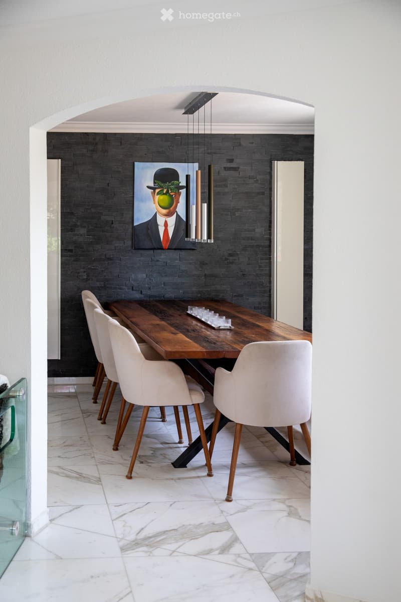 Dining room with a long wooden table, white chairs, a modern pendant light fixture, and a painting on a dark brick wall.