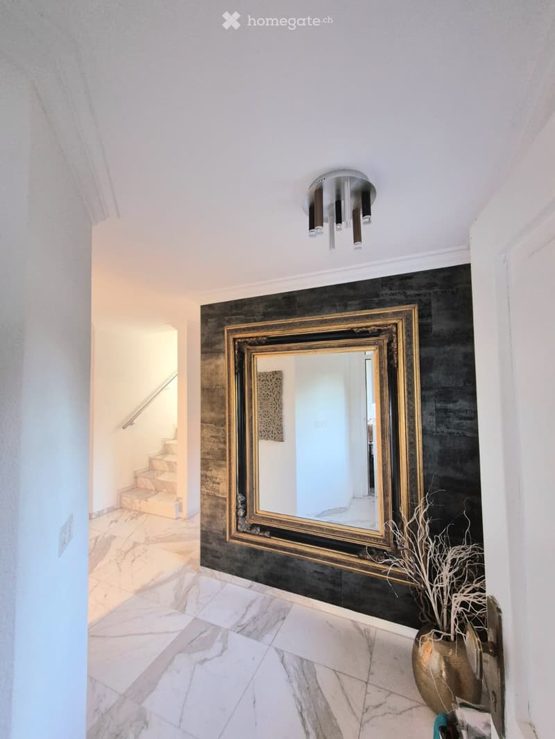 Elegant hallway with white marble flooring, a large gold-framed mirror on a dark accent wall, and a decorative gold vase with dried branches. A modern chandelier hangs from the ceiling, and a staircase leads upward.