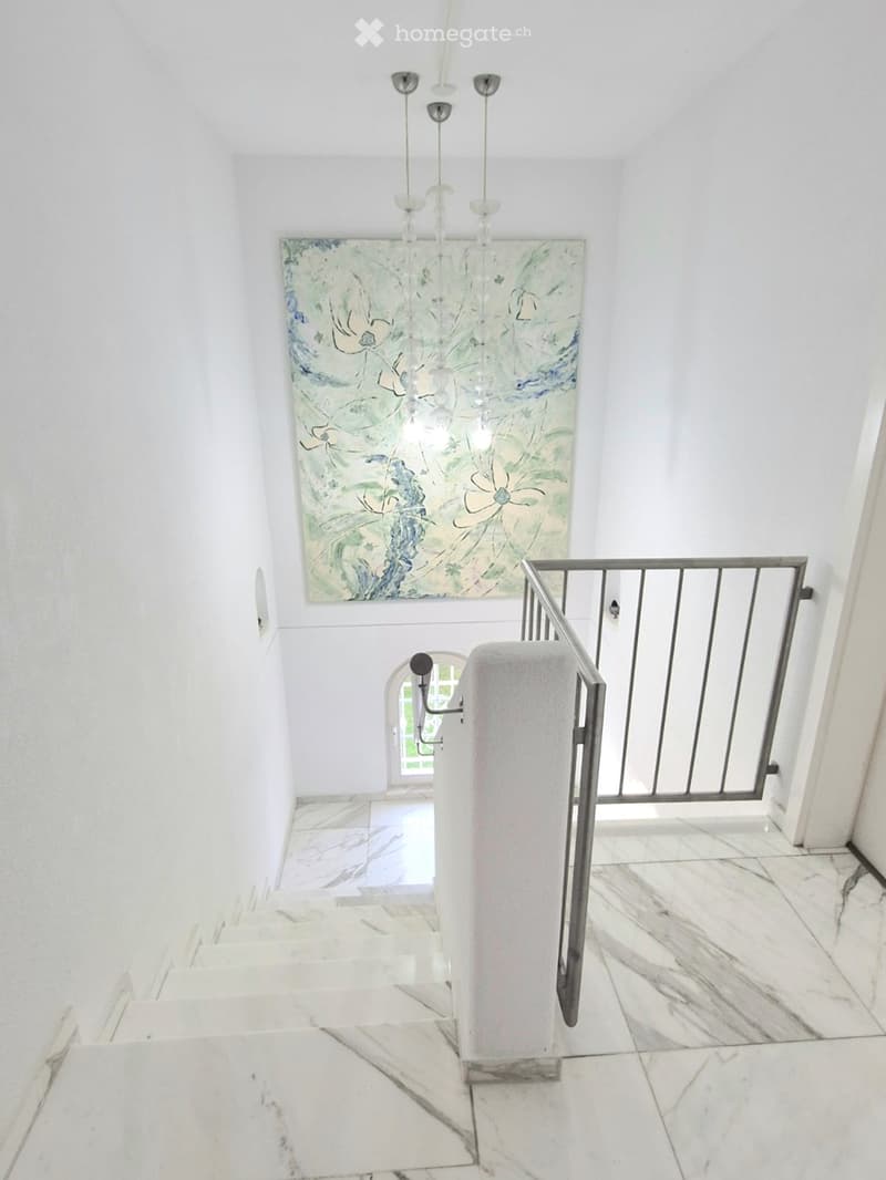 Staircase with white marble steps, modern metal railing, and a large abstract painting on the wall above. The hallway has white walls and a chandelier hanging from the ceiling.