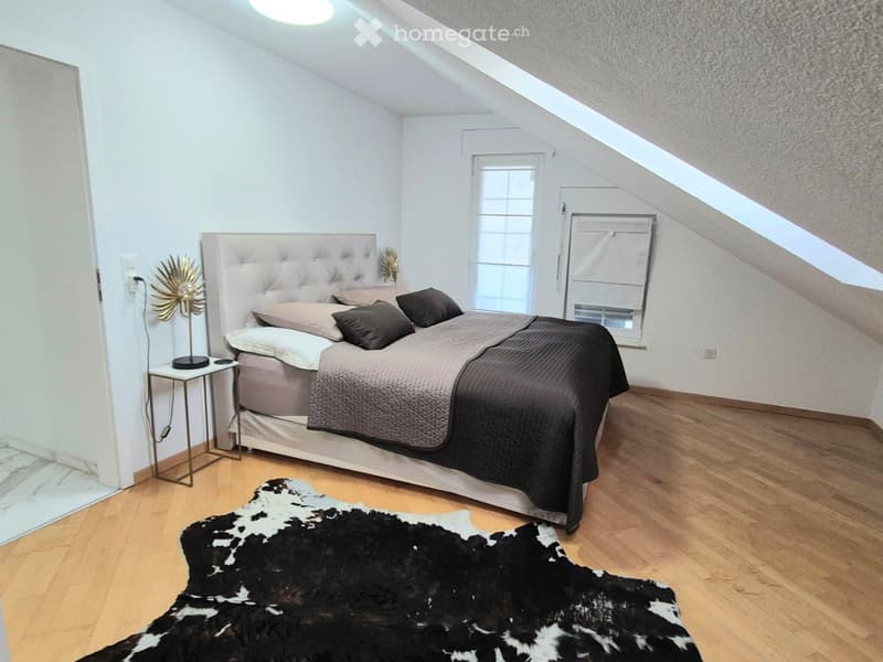Cozy attic bedroom with wooden floors, slanted ceilings, large window, upholstered headboard, nightstand, and decorative lamp.