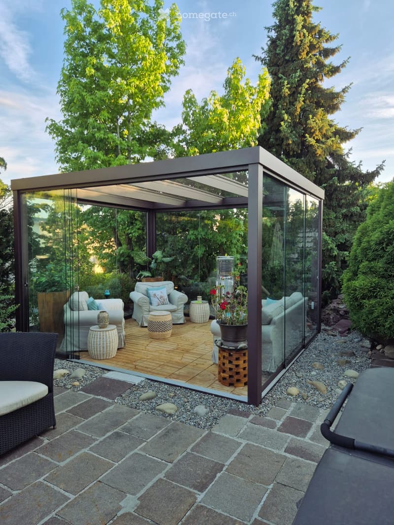 A modern glass-enclosed patio with wooden flooring, featuring a sofa, chairs, tables, and potted plants, surrounded by a stone-paved garden area with trees.