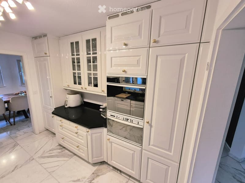 Modern kitchen with white cabinets, black countertops, and stainless steel appliances including a refrigerator, oven, and dishwasher. A coffee maker is on the counter, and a dining area with a table and chairs is visible in the background.