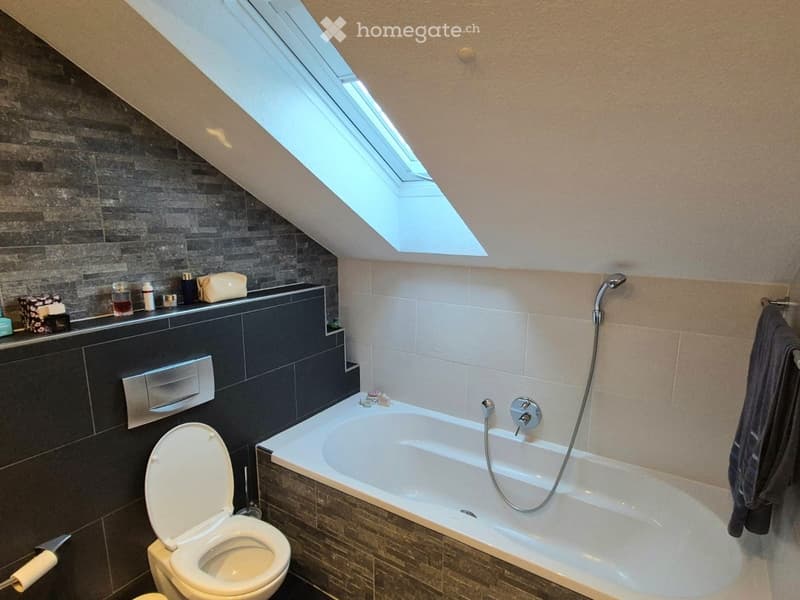 Modern bathroom with a bathtub, toilet, and a skylight window above, featuring dark tiled walls and a towel hanging on the side.