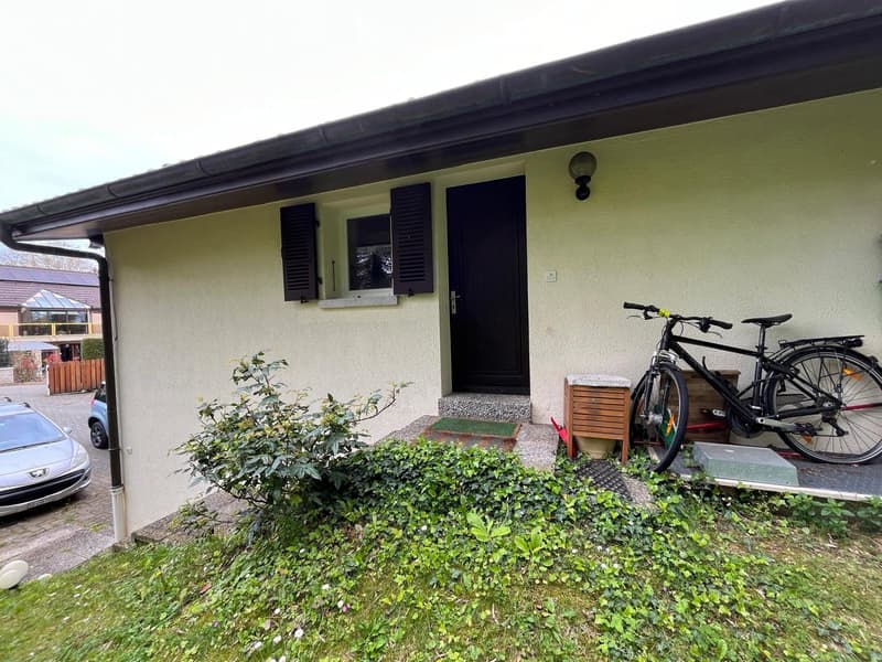 Single-story house with a light green exterior, black front door, small window with shutters, bicycle parked on the porch, and a car parked nearby.