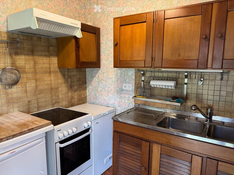 A compact kitchen with wooden cabinets, a white range hood, a Bosch dishwasher, a white oven, a sink with a faucet, and a wooden cutting board on the counter.
