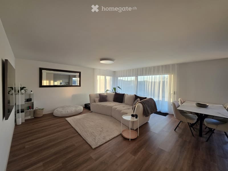 Modern living room with a sectional sofa, coffee table, dining table, chairs, and a large mirror on the wall. The room features wooden flooring, large windows with white curtains, and a cozy rug.