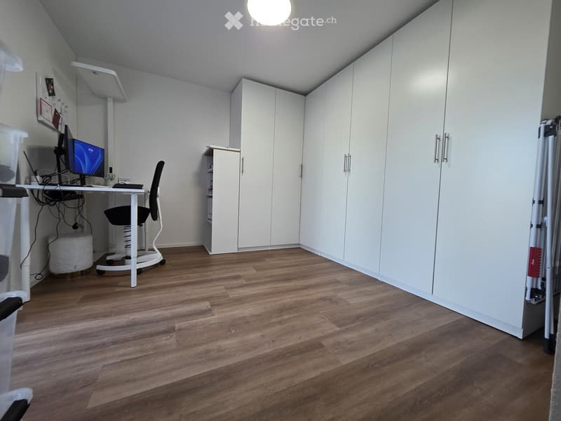A modern, well-lit room with wooden flooring, white walls, and built-in white cabinets. A desk with a computer and chair is positioned against the wall, and a small round stool is nearby.