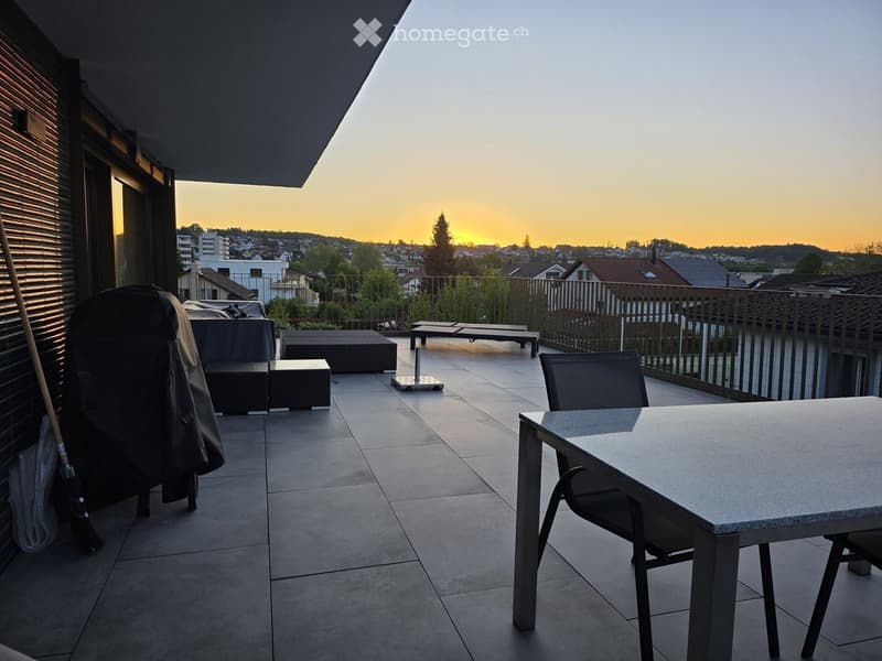 Balcony with tiled floor, outdoor furniture including a table, chairs, sofa, ottoman, and sun loungers, overlooking a cityscape at sunset.