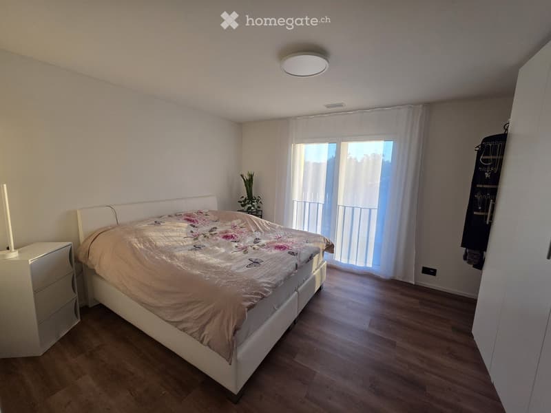 Modern bedroom with a white bed, floral duvet, nightstand, and large window with white curtains. Wooden flooring and a small plant near the window.