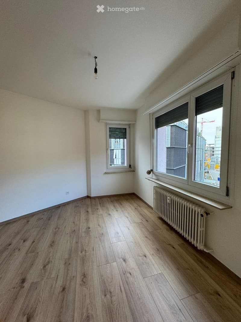 Empty room with light wooden flooring, white walls, and two windows, one with blinds and the other with a radiator below it.