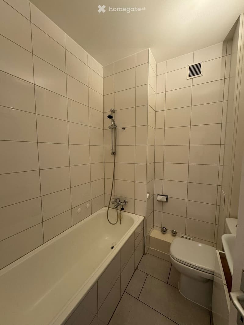 Modern bathroom with a bathtub, toilet, and showerhead. The walls and floor are tiled in a light beige color, and the space is compact with a slanted ceiling.