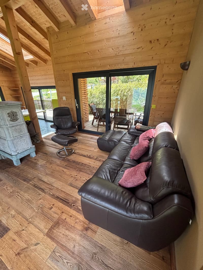 Cozy living room with wooden walls and flooring, featuring a black leather sofa, a black leather chair, a matching stool, and a stone fireplace.