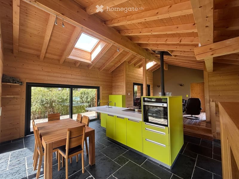 Bright kitchen with wooden beams, green cabinets, a dining table with chairs, and a large window overlooking the garden.