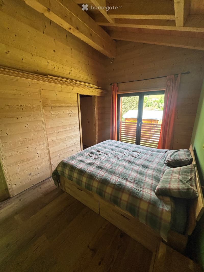 Cozy wooden bedroom with a single bed, plaid bedding, and wooden walls. A window with red curtains offers a view of the outdoor area.