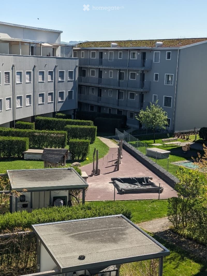 Courtyard view of a multi-story residential building with balconies, surrounded by neatly trimmed hedges, a paved walkway, and a small swimming pool area.