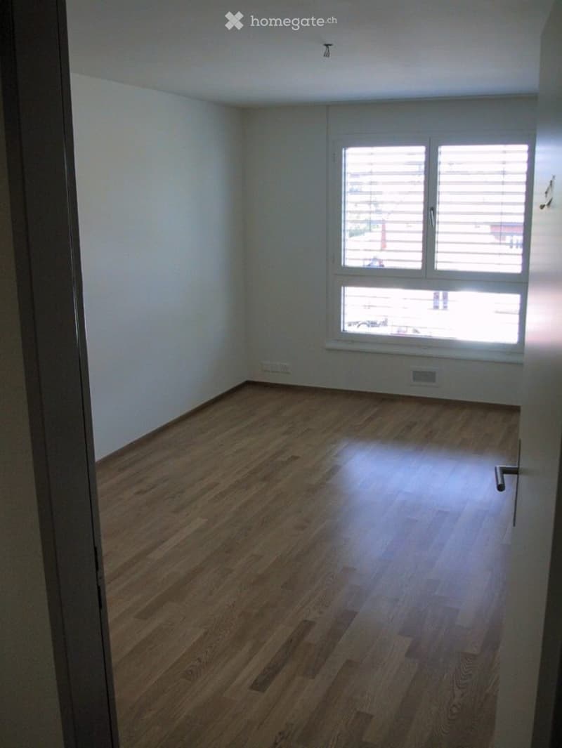 Empty room with wooden flooring and white walls, featuring a large window with blinds.