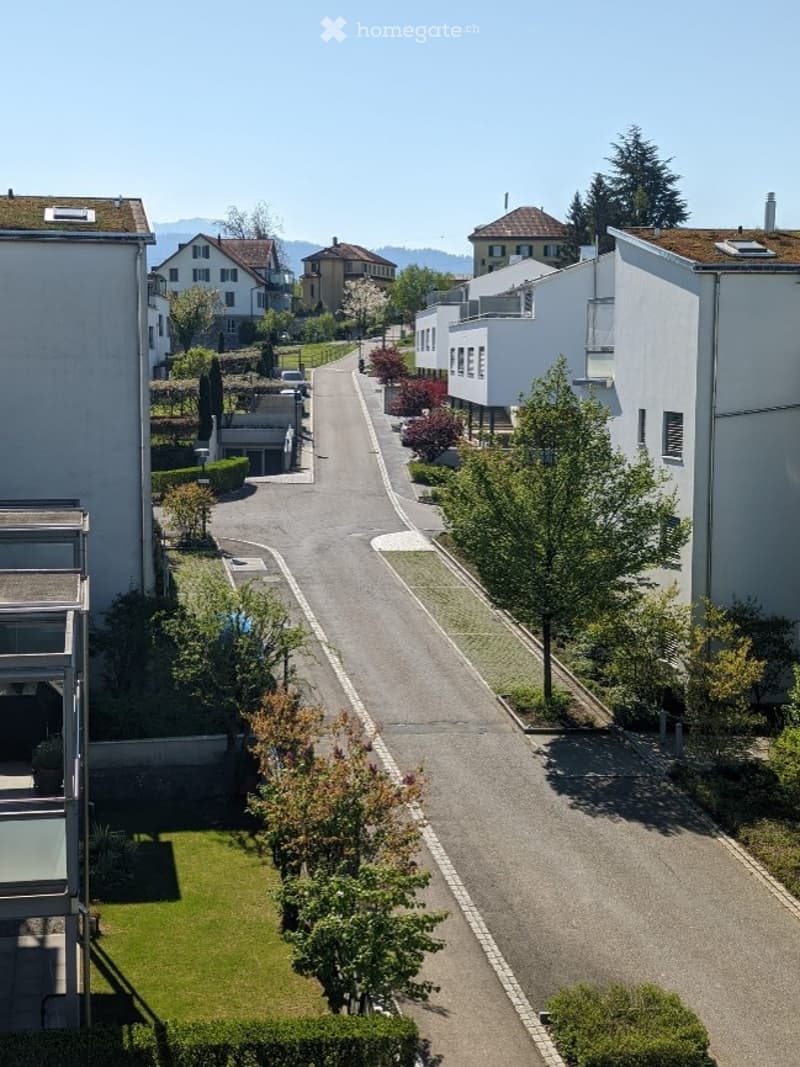 A quiet residential street lined with modern apartment buildings, surrounded by greenery and trees.