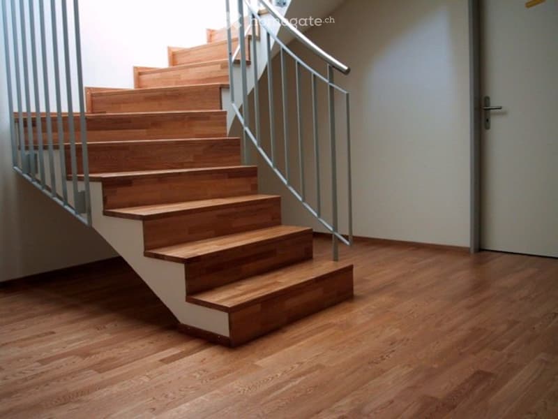 Wooden staircase with metal railing leading up, wooden floor, white walls, and a closed door visible.