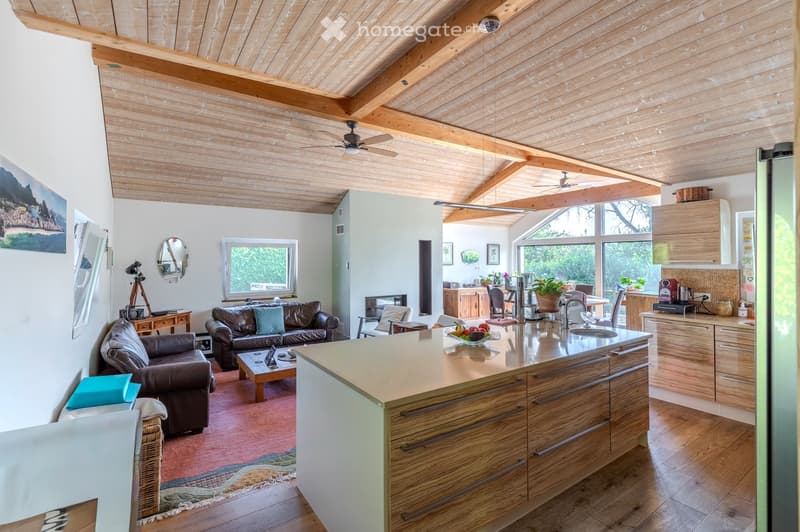 Open-concept living and dining area with a kitchen island, wooden beams, and large windows. Features a leather sofa, coffee table, and various kitchen appliances.