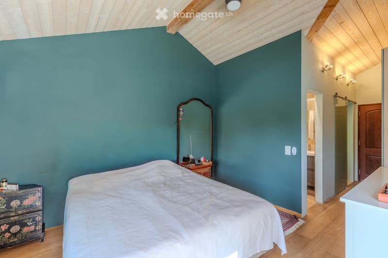 Cozy bedroom with teal walls, wooden floor, and slanted wooden ceiling. Features a large bed with white bedding, a nightstand with a lamp, a decorative drawer, and a mirror on the wall. A small table with items is visible near the bed.