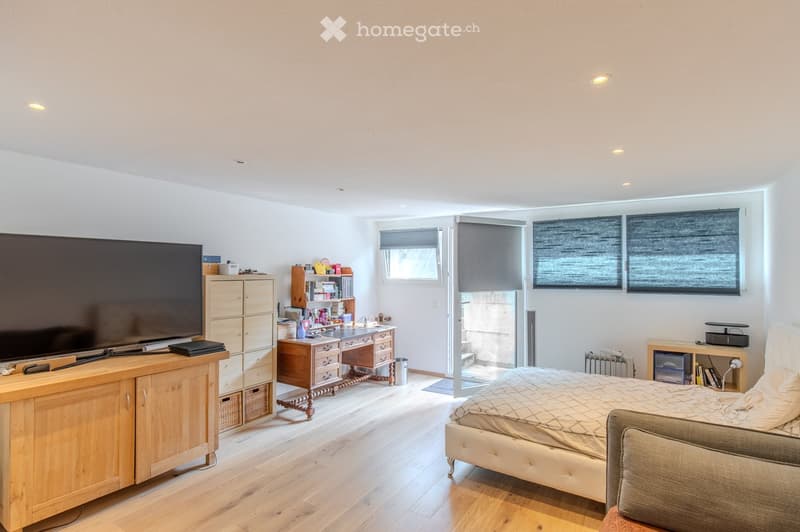 Bright bedroom with wooden flooring, large bed with beige bedding, wooden cabinet, desk with books, and television on a wooden stand.