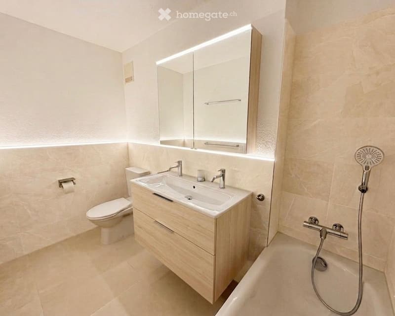 Modern bathroom with a white sink, toilet, mirror, and bathtub. Features a sleek wooden vanity with a built-in sink and a modern showerhead.