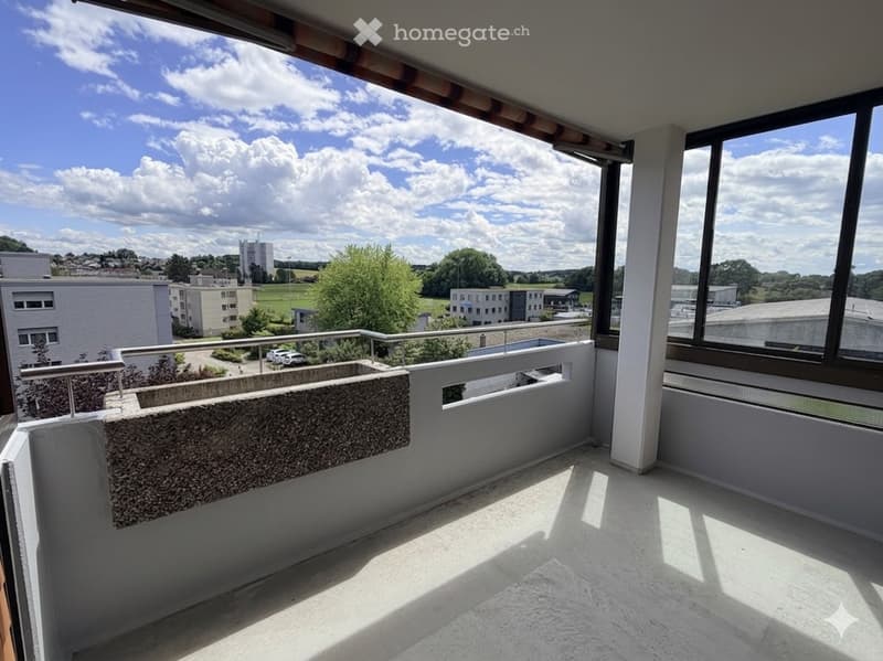 Empty balcony with large windows, metal railing, and a view of the surrounding area with buildings and greenery.
