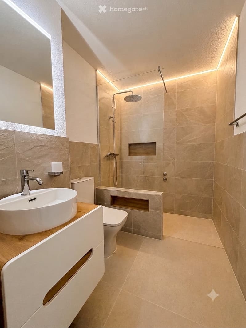 Modern bathroom with a white sink, toilet, and glass-enclosed shower with a rainfall showerhead. Light beige tiles and a sleek wooden vanity.