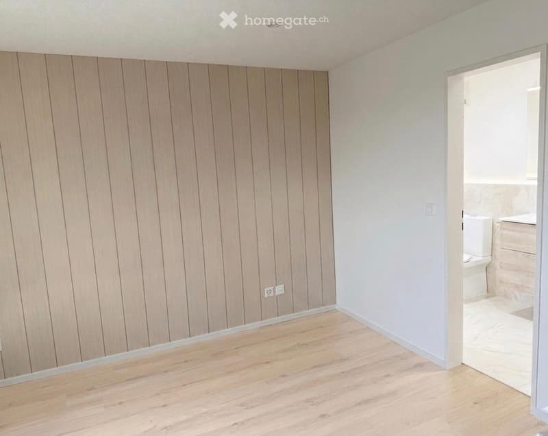 Empty room with wooden flooring and light-colored walls, featuring a partially visible adjacent bathroom with a toilet and sink.