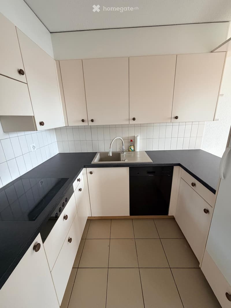 Modern kitchen with white cabinets, black countertops, a sink, faucet, oven, and dishwasher. The floor is tiled, and the walls are tiled with white tiles.
