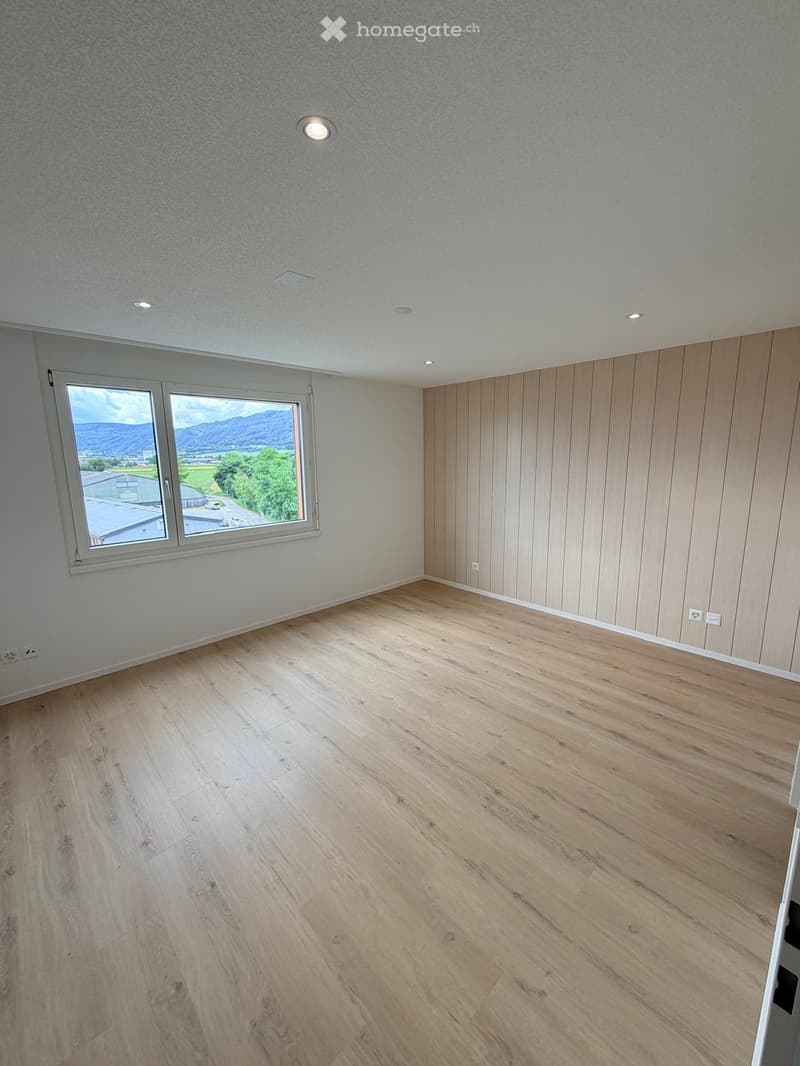Empty room with wooden flooring, white walls, and large windows offering a scenic view of mountains and greenery.