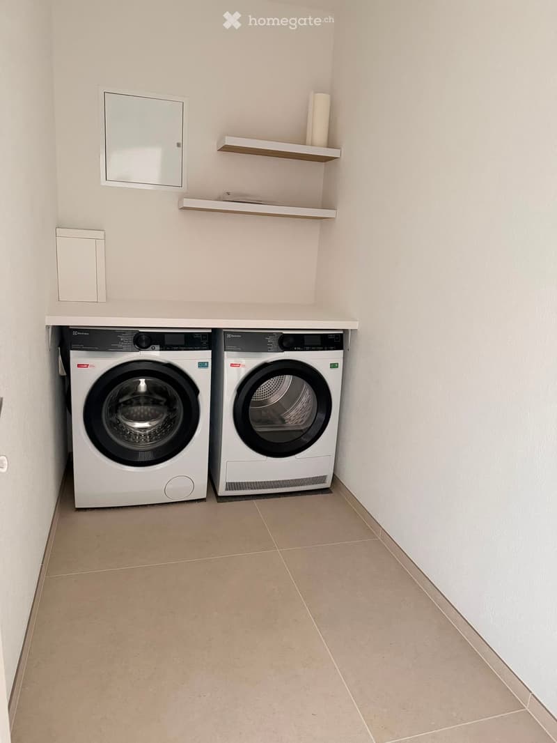 A small, modern bathroom with a washing machine and dryer side by side, a mirror, and a small shelf above the appliances.