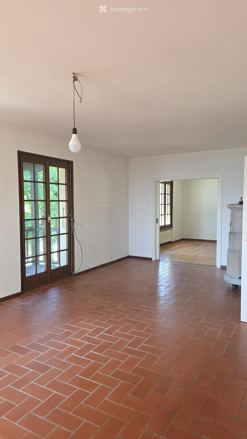 Empty room with red brick tiled floor, white walls, and a large window with glass panels. A hanging light fixture is visible, and an adjacent room is visible through an open doorway.