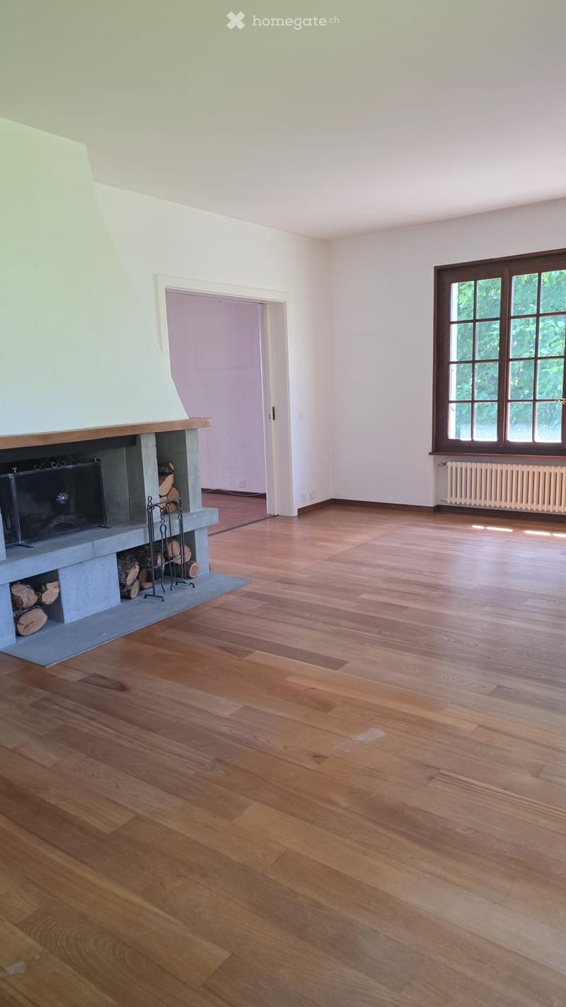 Empty room with wooden flooring, white walls, fireplace with logs, and large windows with brown frames.