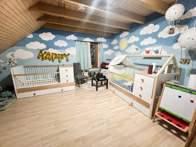 Bright, playful children's bedroom with two cribs, a chair, a small table, and a curtained window. The walls feature a sky-themed mural with clouds and a sun, and the floor is wooden.