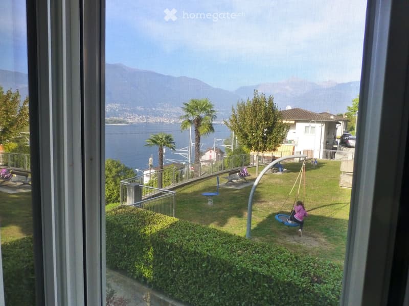 View from a window showing a grassy area with a swing set, a child on a swing, and a scenic lake and mountain view in the background.
